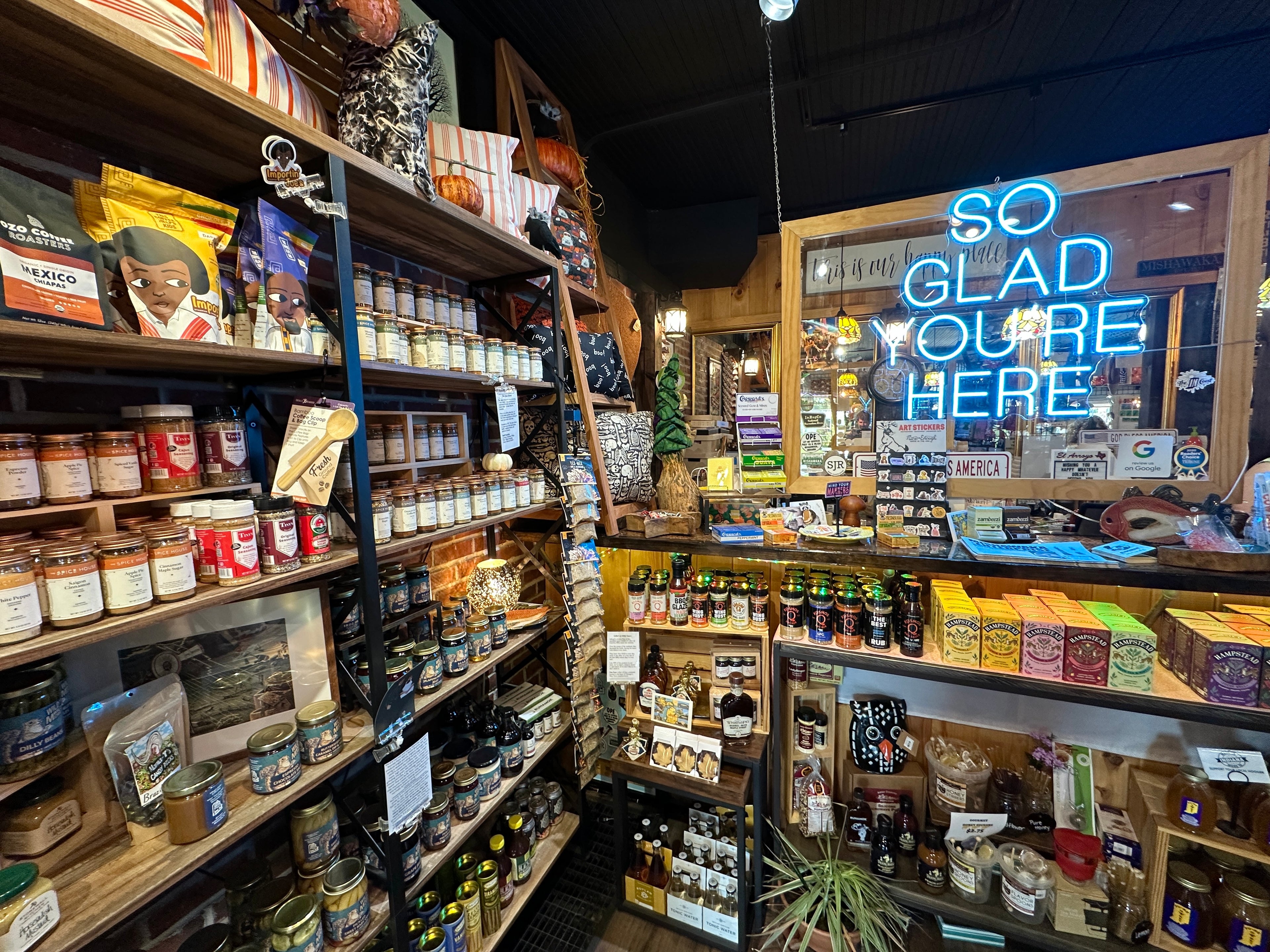 Store interior with shelves stocked with products and a neon sign reading 'So Glad You're Here'.